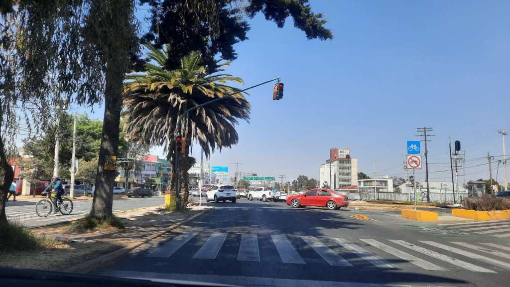 Vista de una intersección en Toluca, con semáforo en rojo y un ciclista en la acera. Se observan vehículos en circulación y edificios al fondo.