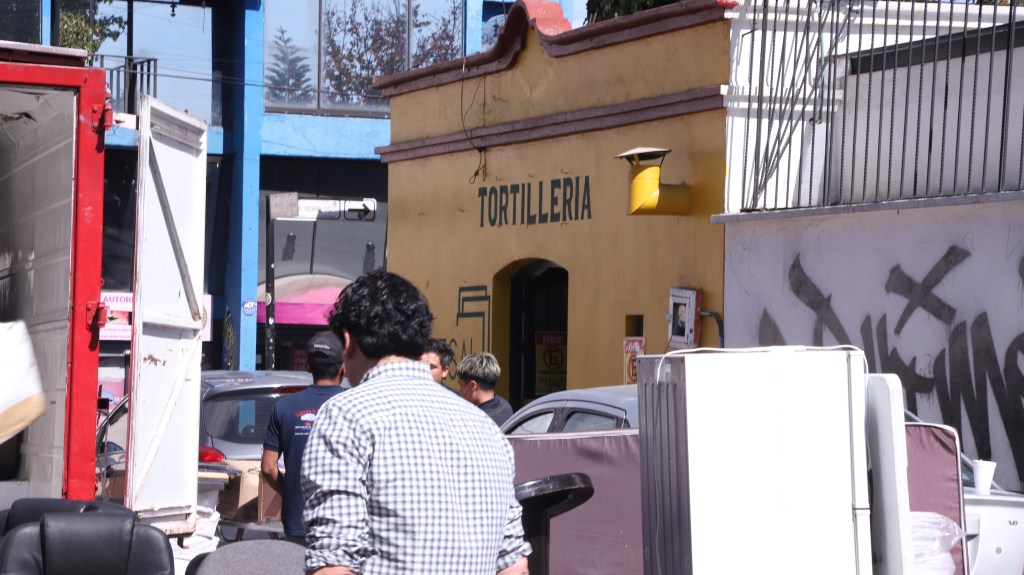 A busy street scene featuring a tortilleria storefront in a yellow building, with people walking and parked cars nearby.