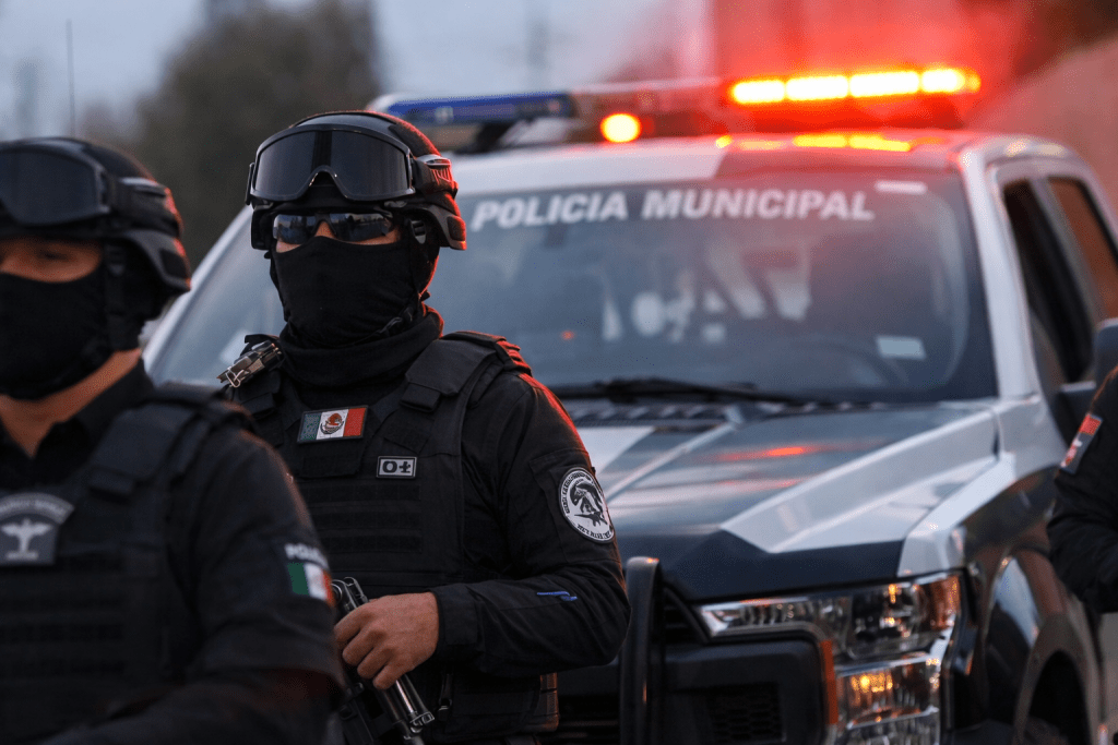 Group of armed police officers in tactical gear with face coverings, standing in front of a police vehicle with emergency lights flashing.