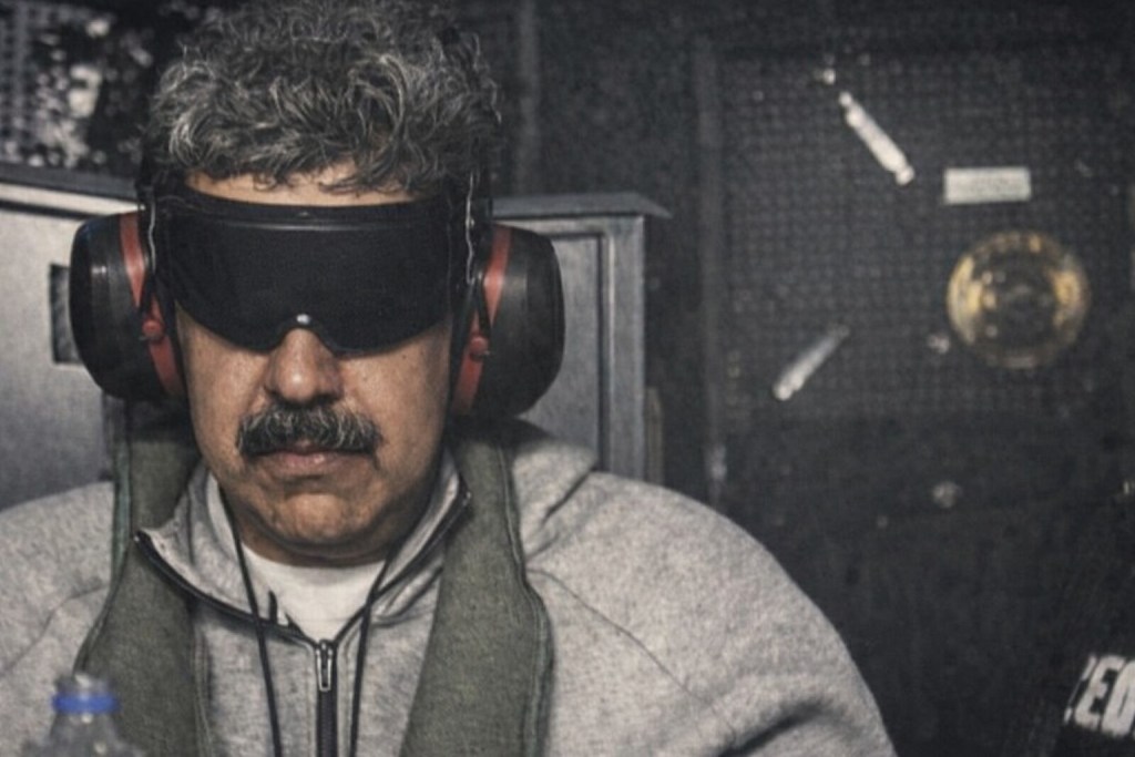 Hombre con gafas de protección y auriculares, sentado frente a un ambiente oscuro con elementos de seguridad.