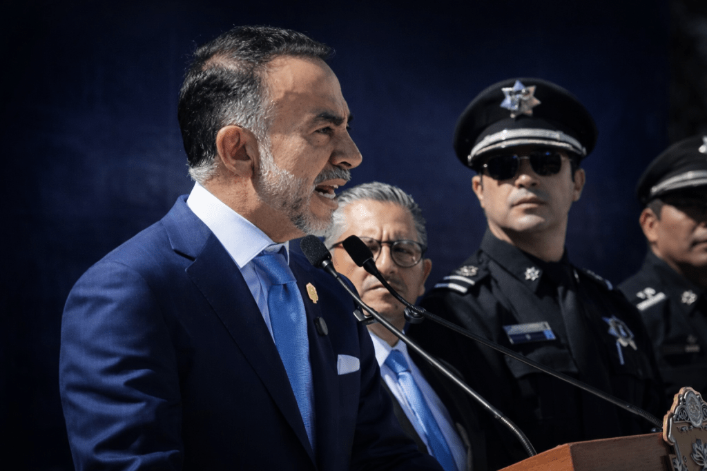 Un hombre en traje azul habla frente a un micrófono durante una conferencia, con dos oficiales de policía al fondo.