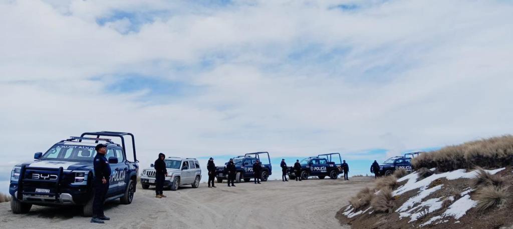 Several police vehicles parked on a dirt road in a mountainous area, with officers standing beside them against a cloudy sky.