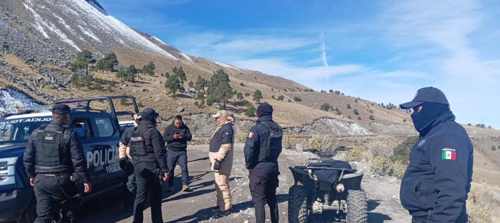 A group of police officers in black uniforms is gathered near a police vehicle and an all-terrain vehicle in a mountainous area with snow on the road and a clear blue sky.