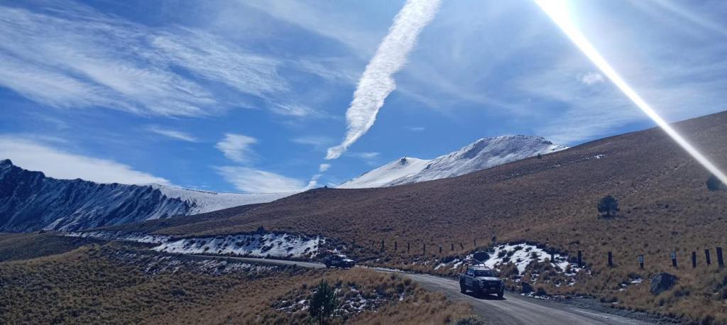 A scenic view of a mountainous landscape with snow-covered peaks, a dirt road winding through grassy terrain, and two vehicles parked along the road under a blue sky with wispy clouds.