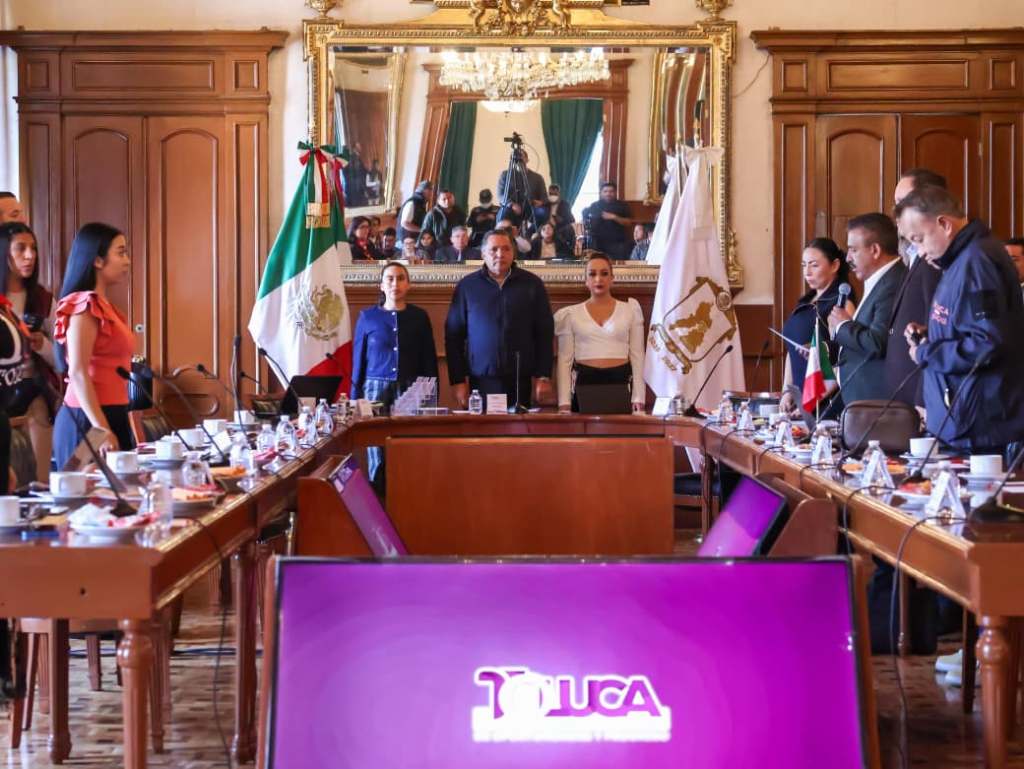 A formal meeting in a government setting with officials standing and seated around a conference table, featuring the Mexican flag and a large mirror reflecting the event.