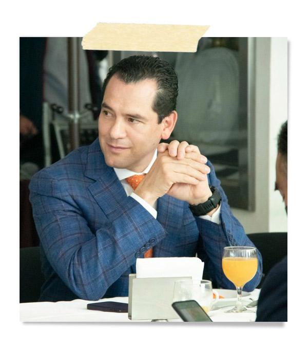 Hombre sentado en una mesa, vestido con un traje a cuadros azul y corbata naranja, mirando hacia un lado mientras se apoya en sus manos. Hay vasos y un menú en la mesa.
