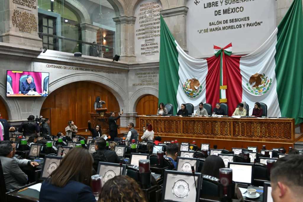 A view of the Mexican Congress chamber during a session, with officials seated at the front and a large screen displaying a speaker. The chamber features decorative flags and ornate wooden accents.