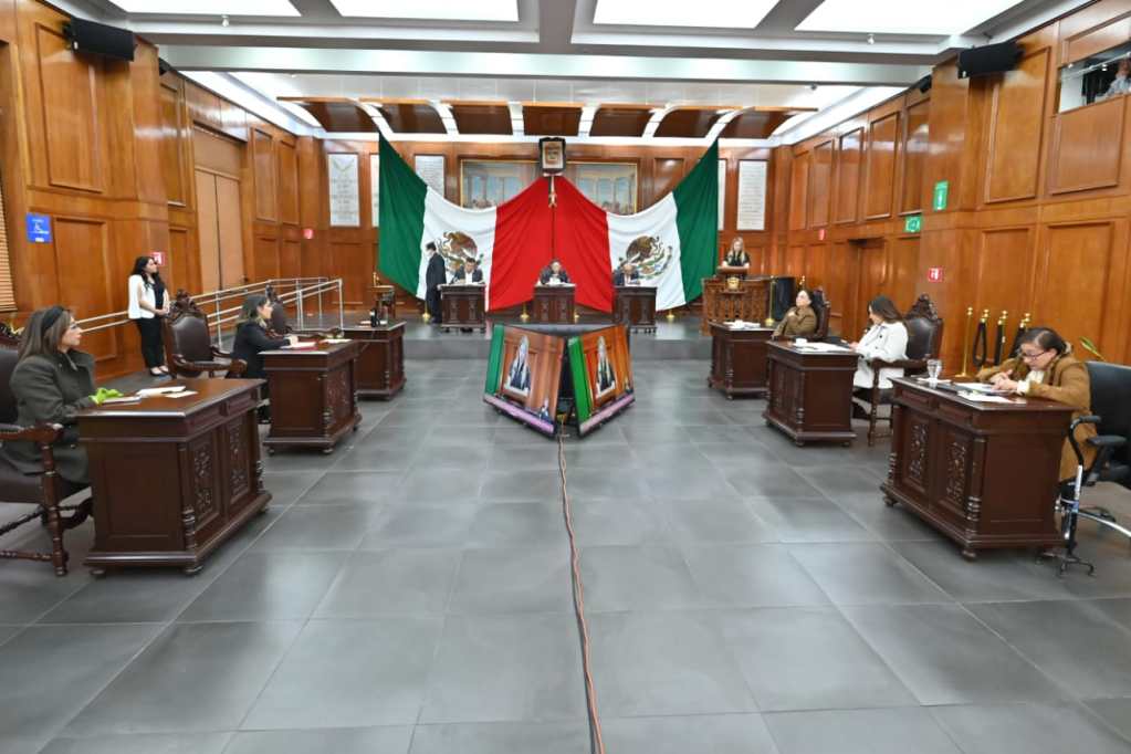 A legislative chamber with wooden walls and furniture, featuring several people seated at desks. The Mexican flag is displayed prominently in the background.