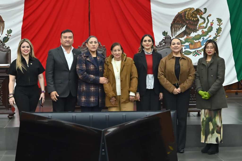 Group of seven individuals standing together in front of the Mexican flag, with prominent colors of red, white, and green in the background.
