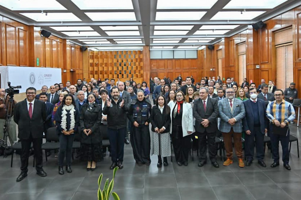A diverse group of people standing together in a conference room, many smiling and some applauding, with wooden paneling in the background and seated attendees visible.