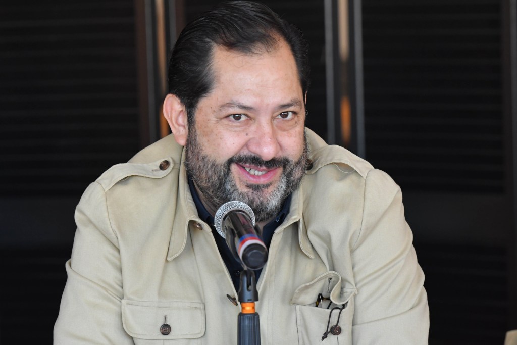 A man with a beard smiling while speaking into a microphone, wearing a light-colored jacket.