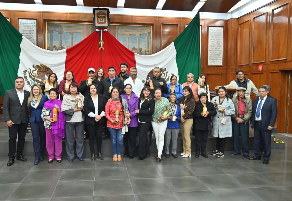 Group photo of individuals holding bottles, standing in front of a large Mexican flag backdrop in a formal setting.