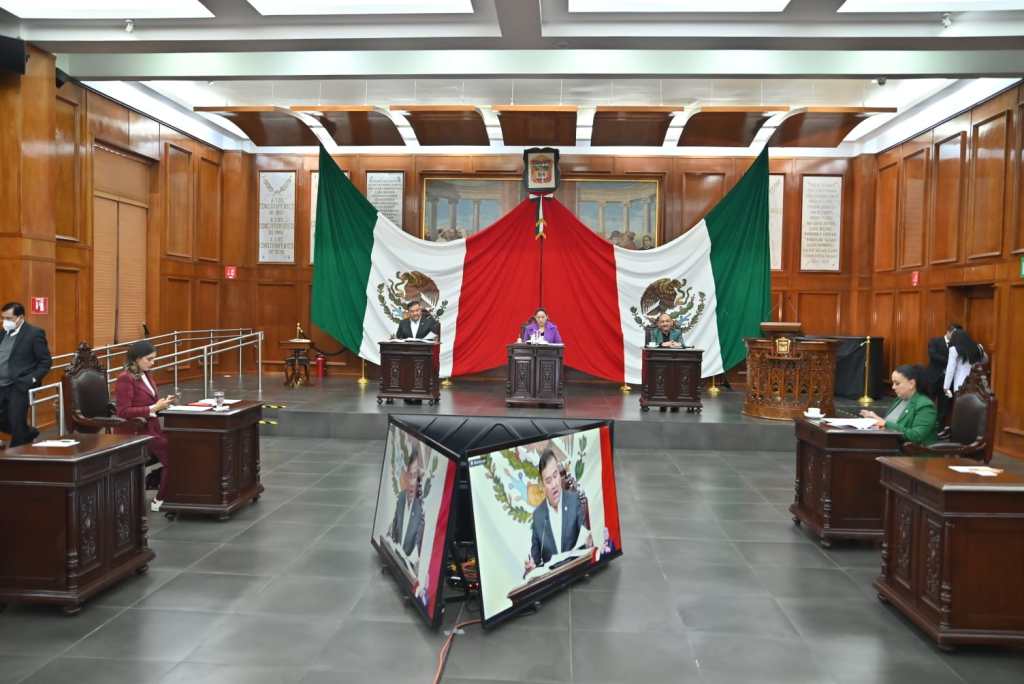 A legislative assembly session in Mexico, featuring multiple officials seated at desks, with large Mexican flags displayed in the background.