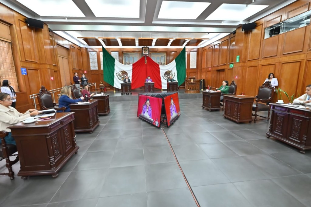 Interior view of a legislative assembly room with wooden walls and traditional decor, featuring the Mexican flag prominently displayed in the background. Several officials are seated at desks, engaged in discussions, while two digital displays are set up in the center.