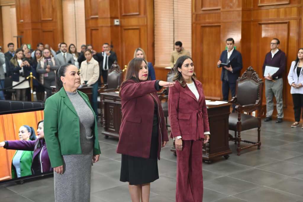 Three women standing with raised right hands during an official ceremony in a formal setting, surrounded by an audience. One woman is wearing a green outfit, another in a red suit, and the third in a pink suit.