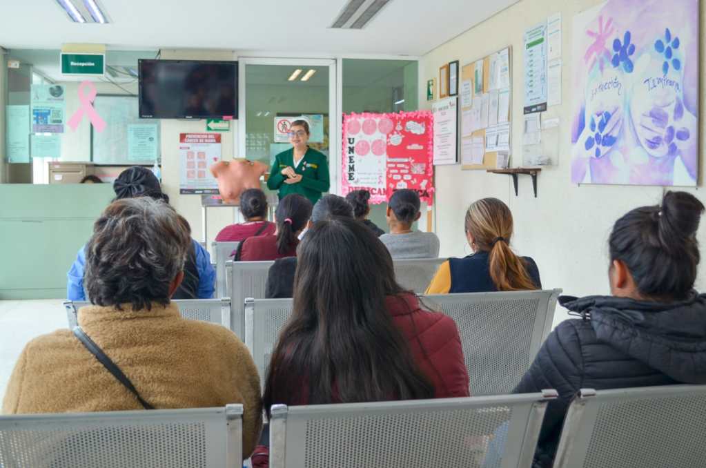 Grupo de mujeres sentadas en una sala esperando para una presentación, con una instructora hablando frente a un cartel informativo.