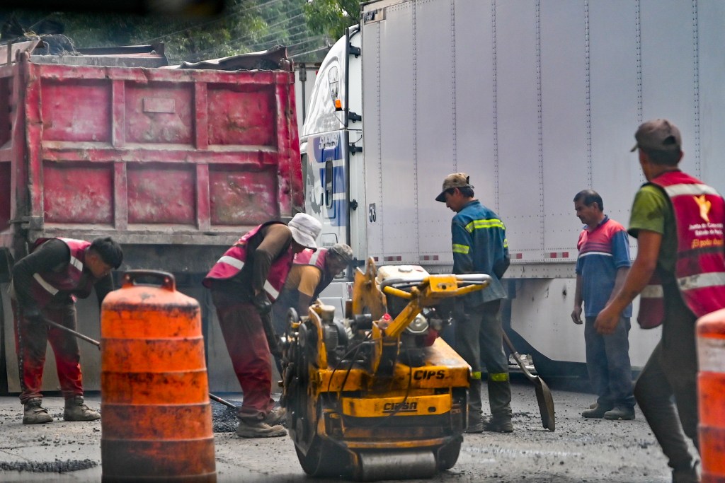 Trabajadores de la construcción realizando labores en la calle, con maquinaria y vehículos de obras en el fondo.