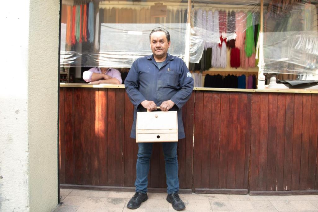 Hombre sosteniendo una caja de madera frente a un establecimiento con telas de colores en el fondo.