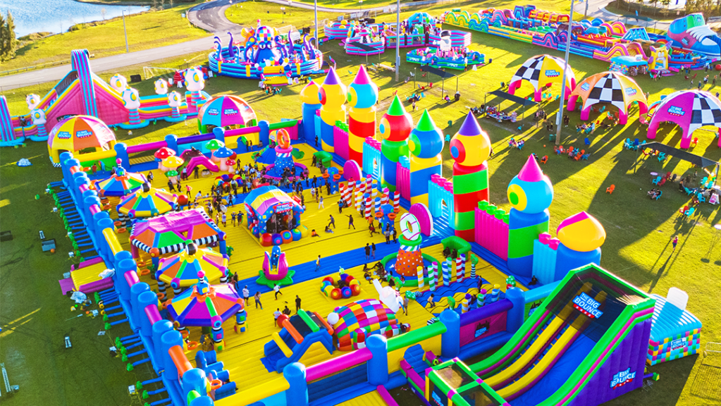 Vista aérea de un parque inflable colorido con múltiples áreas de juegos, toboganes y estructuras interactivas, lleno de niños y familias disfrutando del espacio.