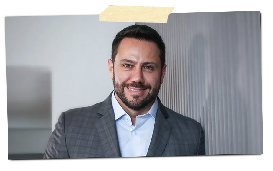 Retrato de un hombre sonriendo, vestido con un traje gris y camisa blanca, de pie frente a una pared con textura.
