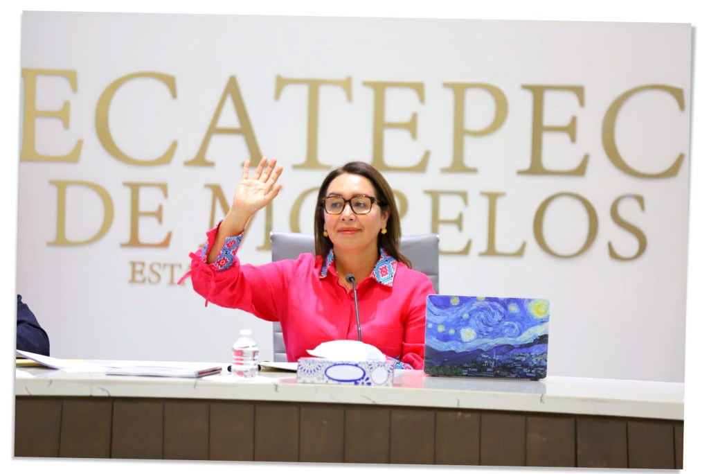 Funcionaria de Ecatepec levantando la mano en señal de saludo, sentada frente a una mesa con una laptop y decoraciones en un entorno institucional.