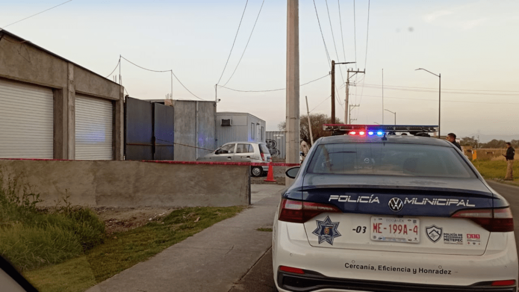 Patrulla de policía municipal con luces encendidas en la carretera, frente a un establecimiento marcado con cinta de seguridad.
