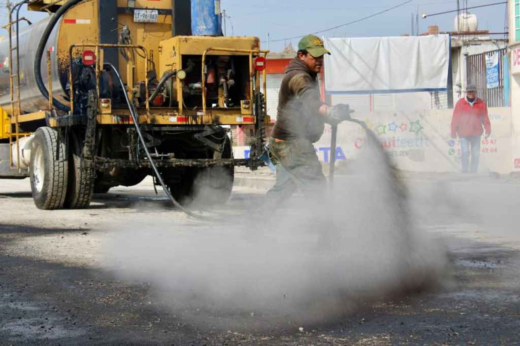 A worker operating a hose to spray a liquid material on a street, with a large yellow truck in the background, and another person walking in the distance.