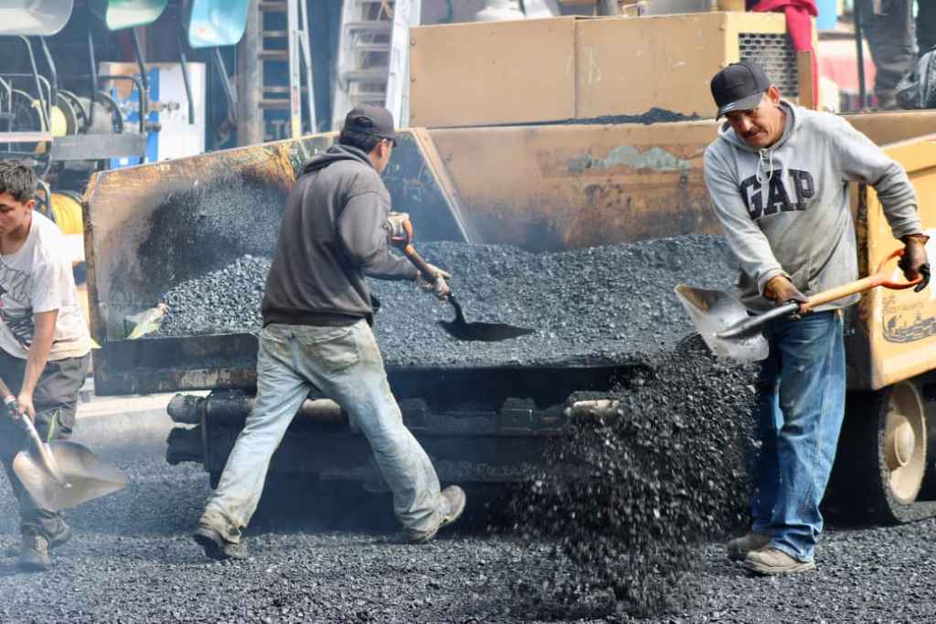 Workers shoveling asphalt from a truck onto a construction site, with machinery in the background.