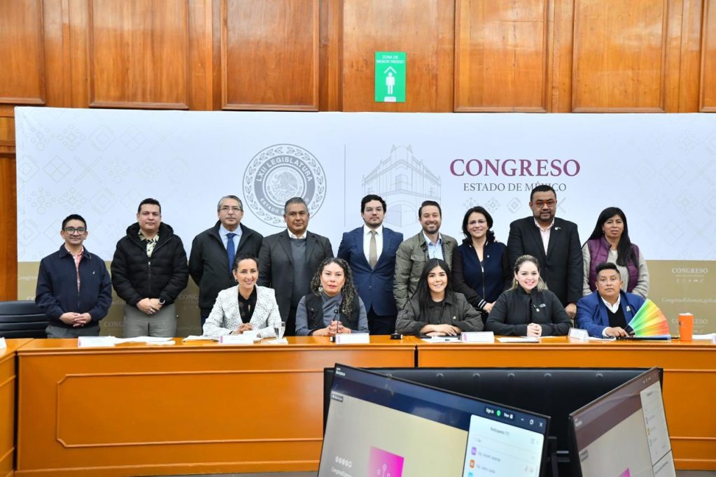 A group of twelve individuals posing for a formal photo in a legislative assembly setting, with a banner in the background that reads 'Congreso Estado de México'. The group includes both men and women, some sitting at a long table and others standing behind.