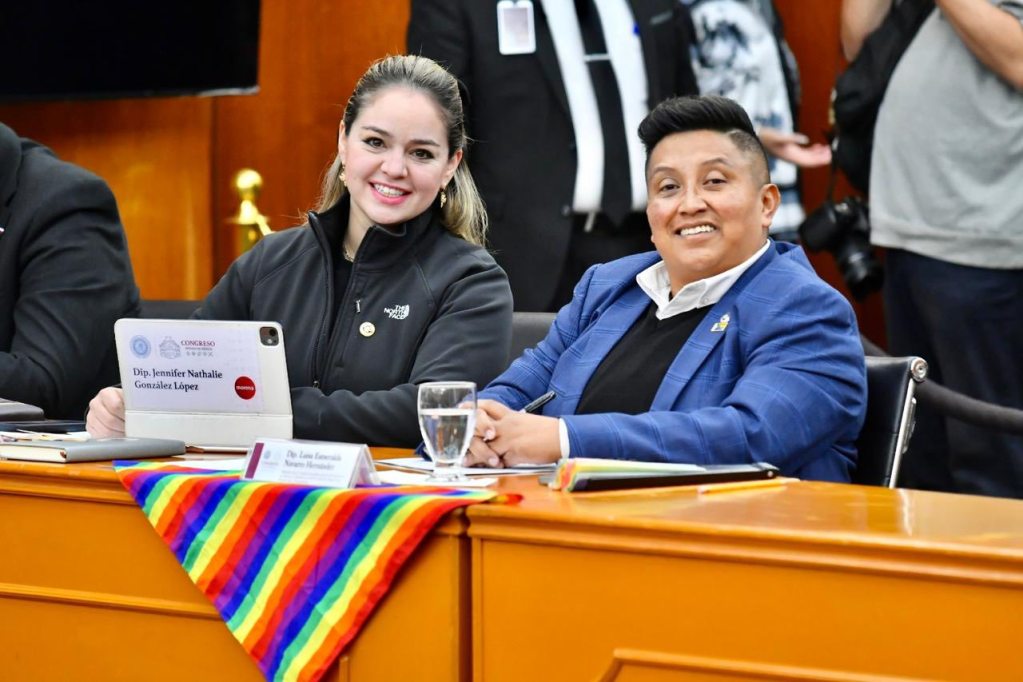 Two individuals smiling at a meeting table, one holding a tablet with their name displayed, while a colorful rainbow-striped cloth is draped across the table.