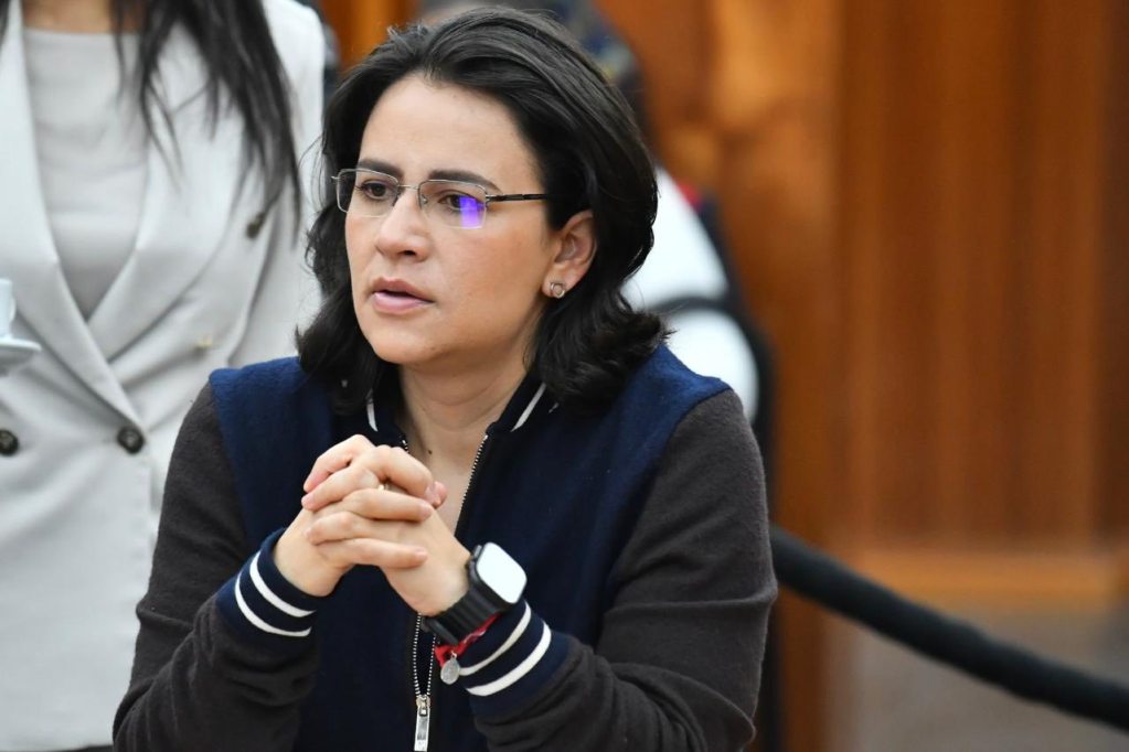 A woman with short black hair and glasses is sitting at a table, looking thoughtfully with her hands clasped together. She is wearing a dark sweater with a blue collar and appears to be engaged in a discussion.