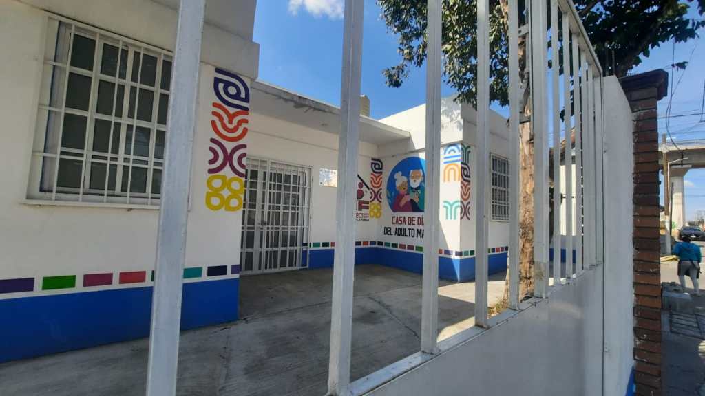 Exterior view of a brightly colored building with a sign that reads 'Casa de Día del Adulto Mayor' visible through a fence.