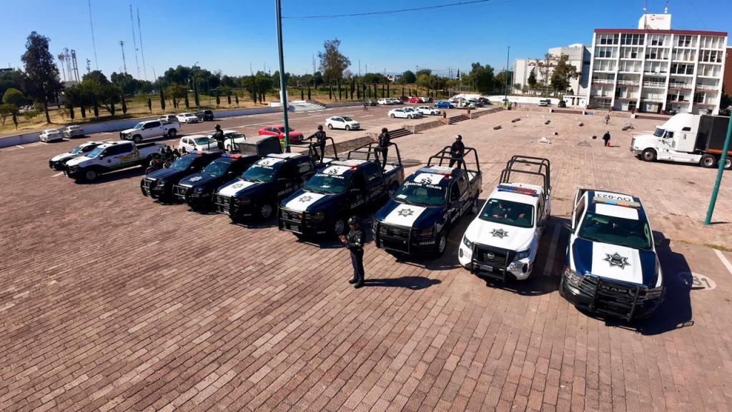 A lineup of police vehicles parked in a spacious area, with officers standing by the cars. In the background, there are additional vehicles and a building.