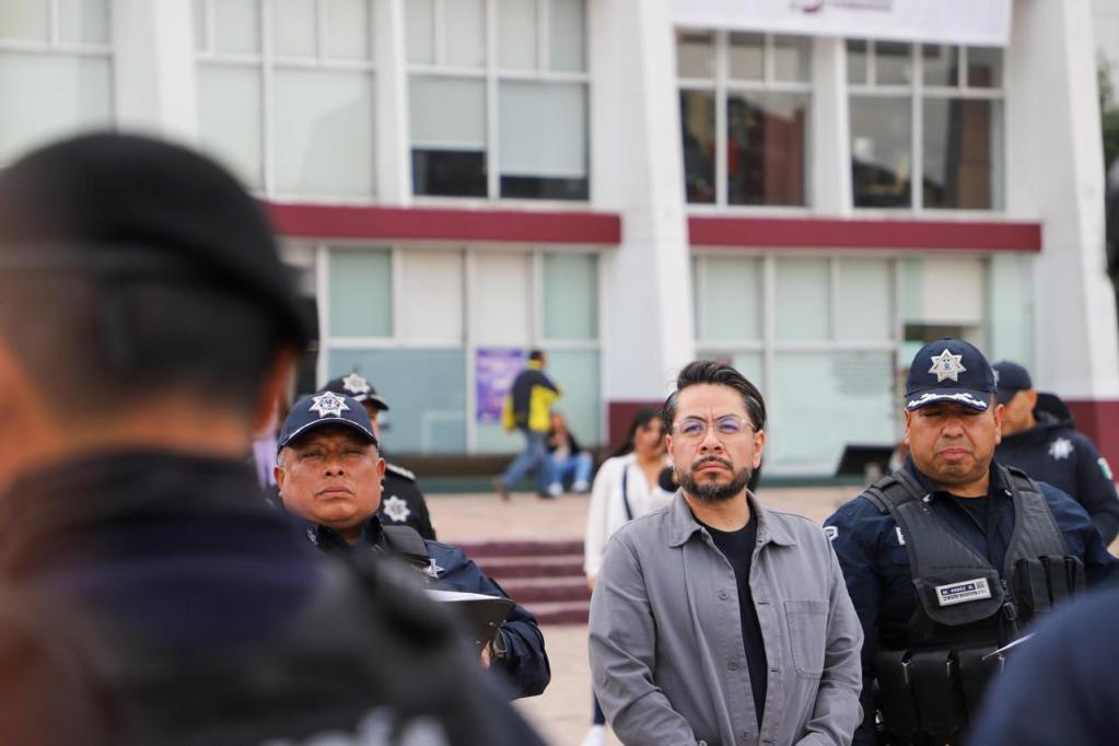 A man with glasses stands in front of police officers outside a building, with several people visible in the background.
