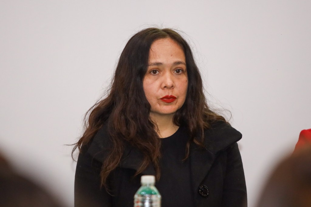 A woman with long, wavy dark hair and red lipstick is seated at a conference table, wearing a black coat and looking serious.
