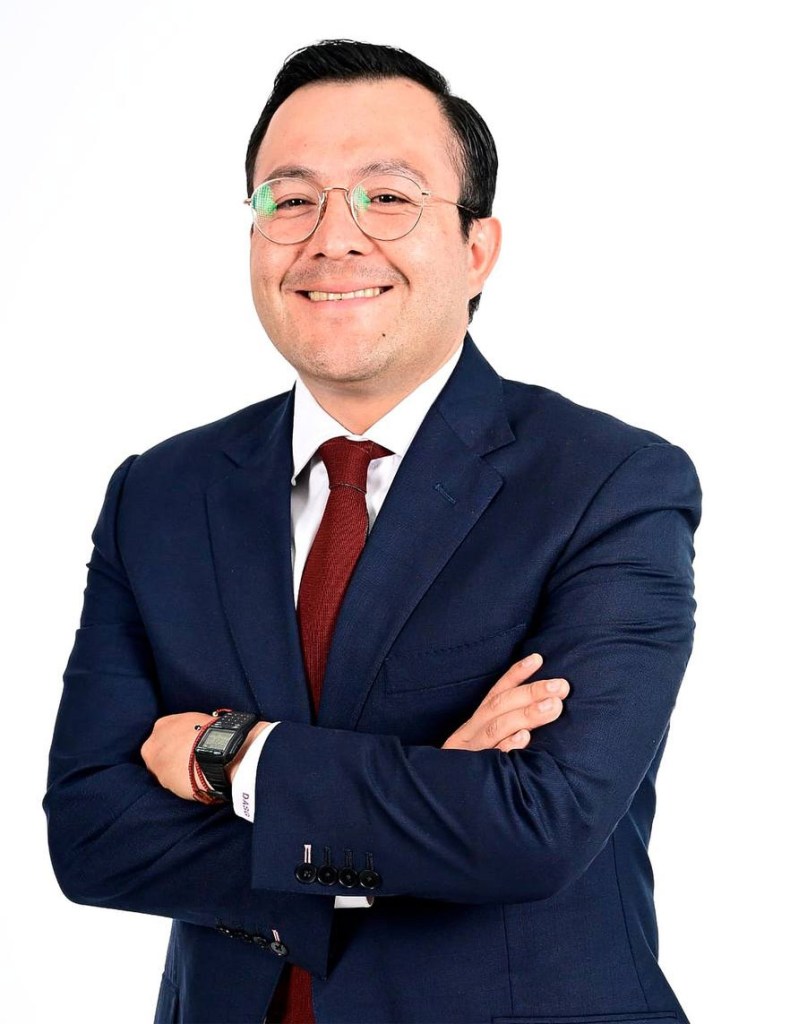Retrato de un hombre vestido con traje oscuro y corbata, sonriendo y con los brazos cruzados frente a un fondo blanco.