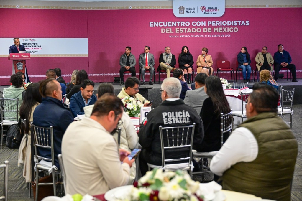 A press conference in Toluca, State of Mexico, featuring a speaker at a podium and several seated panelists, with an audience of journalists and attendees.