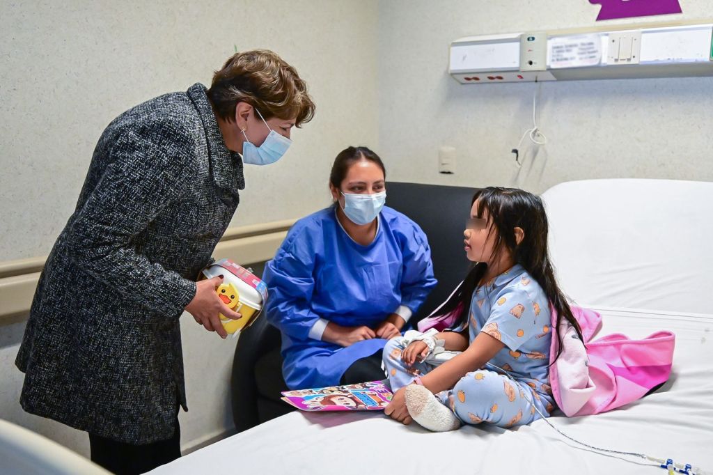 La gobernadora Delfina Gómez Álvarez entrega juguetes a una niña en un hospital, acompañada por una enfermera.