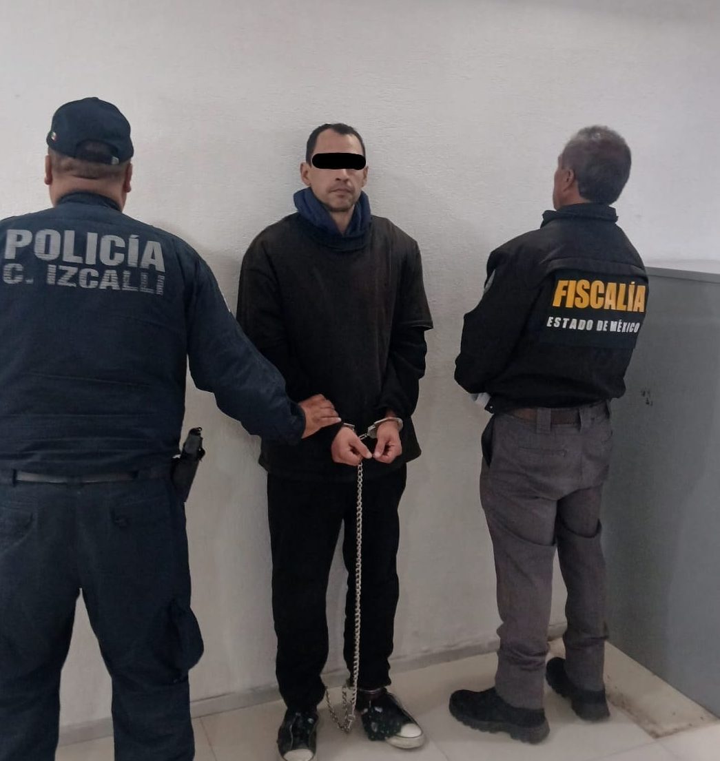A man in handcuffs stands between two police officers in a room. One officer is in a dark blue uniform labeled 'POLICÍA' while the other wears a jacket labeled 'FISCALÍA ESTADO DE MÉXICO'.