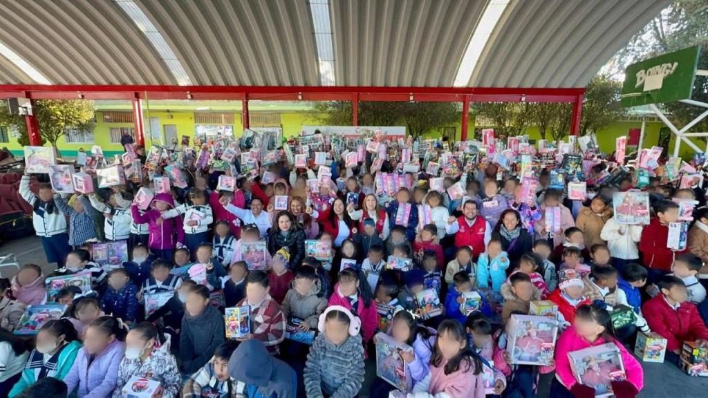 A large group of children and adults gathered under a covered area, holding up toys and gifts, with many children smiling and celebrating.