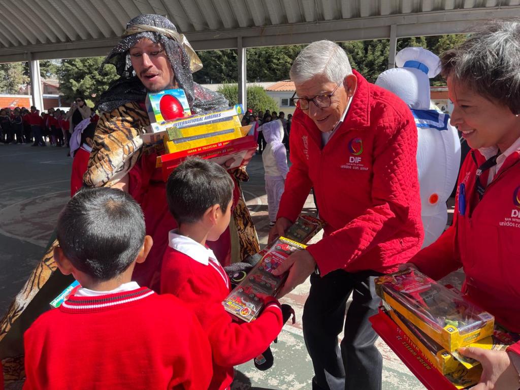 A group of children in red sweaters receive toys from adults in red jackets during an event. One adult is dressed in a colorful costume, while another adult is handing toys to the children.
