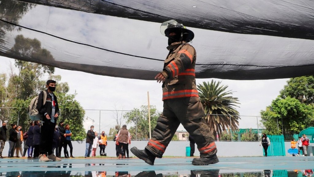 Bombero caminando bajo una estructura de seguridad mientras se desarrolla un simulacro de evacuación, con personas observando al fondo en un espacio al aire libre.