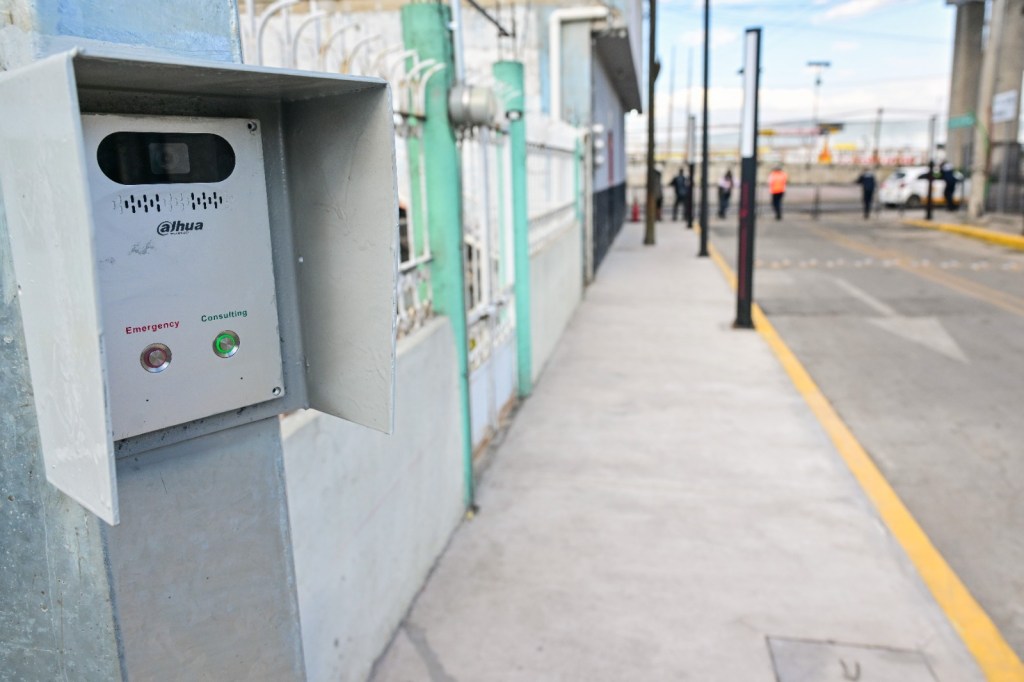 A close-up of a communication device mounted on a wall, featuring buttons labeled 'Emergency' and 'Consulting.' The image shows a sidewalk with people in the background.