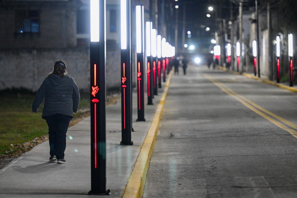 A woman walking along a dimly lit street lined with tall light poles featuring bright white and red accents at night.