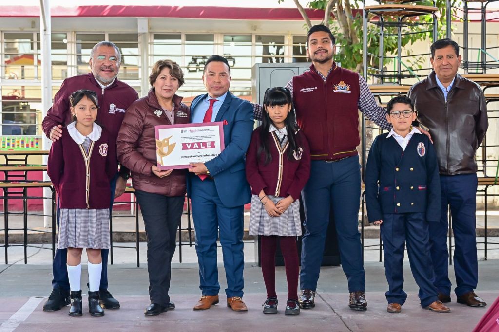 A group of adults and school children standing together in an outdoor setting, holding an educational infrastructure award. The adults are wearing jackets with logos, and the children are in school uniforms.