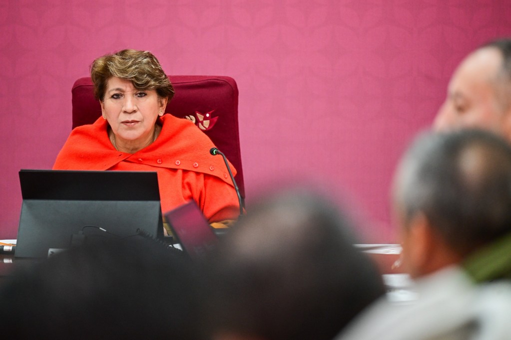 Una mujer con cabello corto, vestida con un abrigo naranja, se sienta ante un micrófono y una computadora, con un fondo de color rosa claro. Se la ve prestando atención a lo que sucede en una reunión.