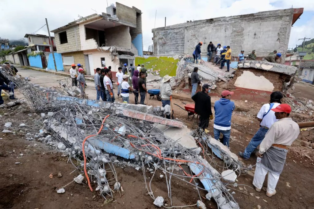Grupo de personas trabajando en la reconstrucción de edificios dañados tras un sismo, con escombros y estructuras colapsadas visibles en la escena.