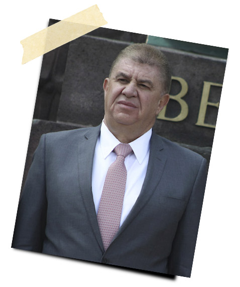 Hombre de pie frente a un edificio, vestido con traje gris y corbata rosa, con expresión seria.