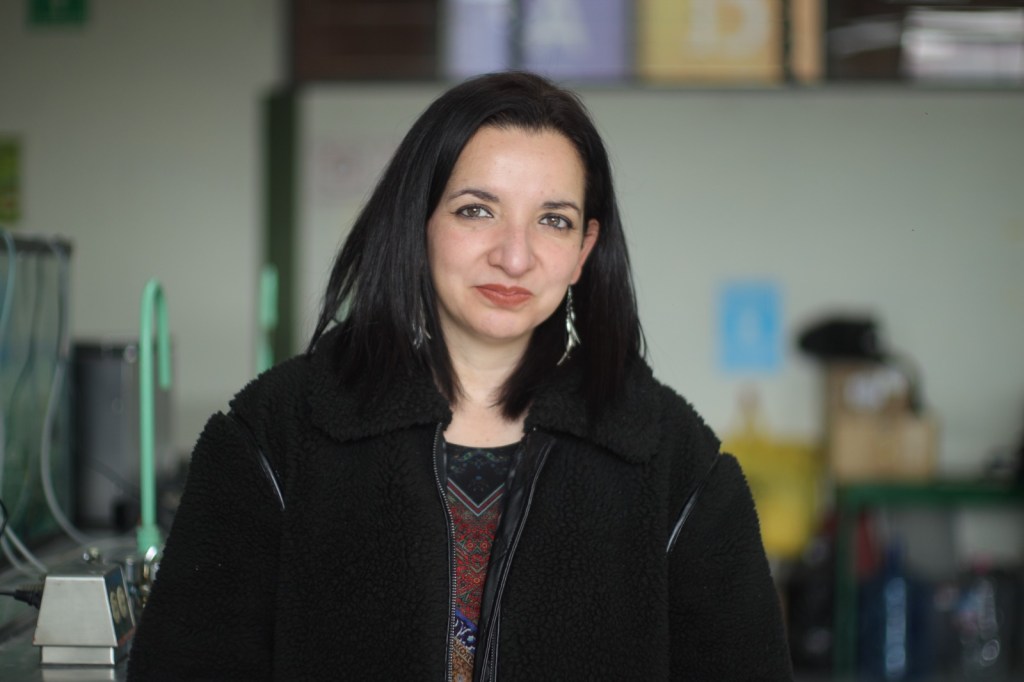 A woman with long black hair wearing a black jacket, standing in a modern indoor space with kitchen equipment and colorful decorations in the background.
