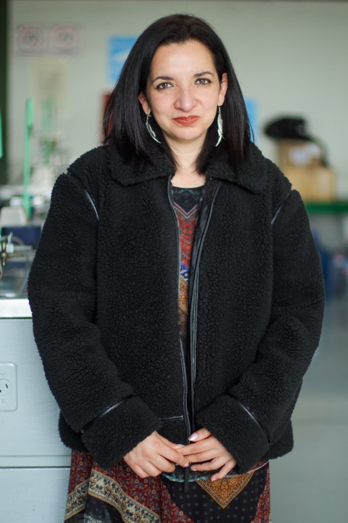 A woman with long dark hair wearing a black fuzzy jacket and large earrings, standing with her hands clasped in front of her, in an indoor space.